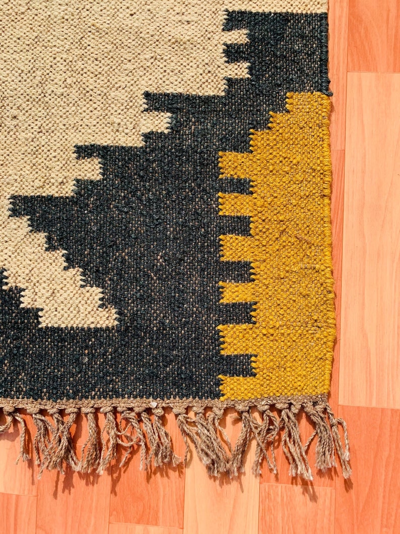 May include: Close-up of a woven rug with a geometric pattern. The rug features a beige background with black and yellow geometric shapes. The rug has a fringe along the bottom edge.