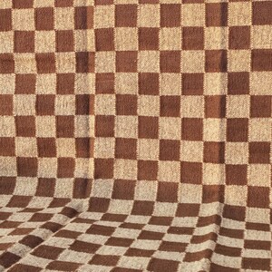 May include: A brown and beige chequered blanket. The blanket has a repeating pattern of large squares in two colours. The fabric appears to be woven, with a slightly textured surface. The blanket is folded, showing the pattern in a three-dimensional perspective.