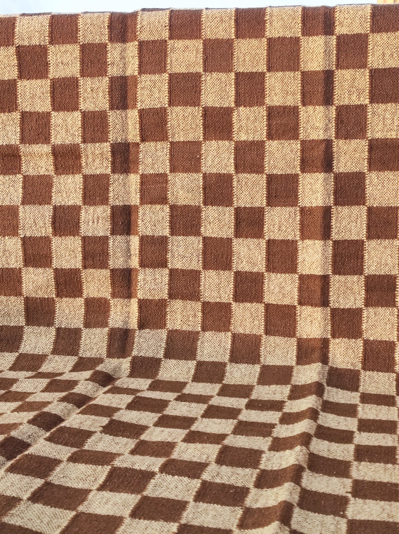 May include: A brown and beige chequered blanket. The blanket has a repeating pattern of large squares in two colours. The fabric appears to be woven, with a slightly textured surface. The blanket is folded, showing the pattern in a three-dimensional perspective.