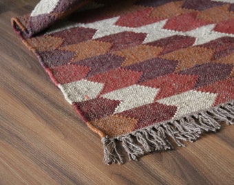 May include: A woven rug with a geometric pattern in shades of red, brown, and beige. The rug has a fringed edge and is partially rolled up, revealing the underside. The rug is placed on a wooden floor.