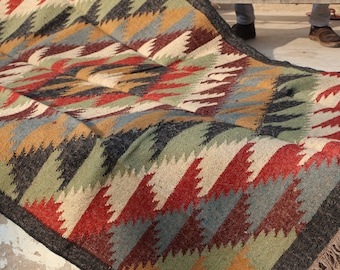 May include: A colorful, woven rug with a geometric pattern. The rug features a repeating design of zigzags in shades of red, green, black, gold, and blue. The rug has a dark border and a fringed edge. The rug is made of a natural fiber.