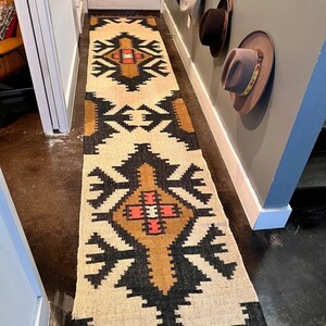 May include: A long, rectangular rug with a geometric pattern in beige, black, brown, and red. The rug is placed in a hallway, with a repeating design of stylized shapes. The rug's design is symmetrical, with a central motif and borders.