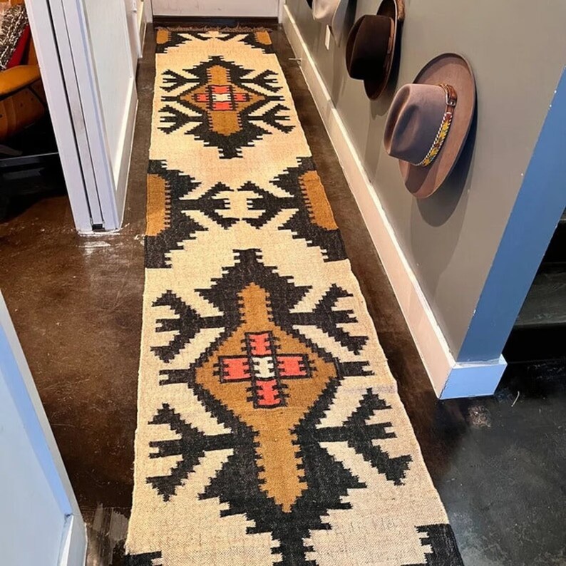 May include: A long, rectangular rug with a geometric pattern in beige, black, brown, and red. The rug is placed in a hallway, with a repeating design of stylized shapes. The rug's design is symmetrical, with a central motif and borders.
