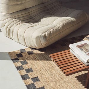 Pu&ograve; includere: Una poltrona bassa beige con tessuto strutturato, vicino a un tappeto tessuto con un motivo a scacchiera nero e beige. &Egrave; visibile anche un tavolino in legno con un libro sopra.