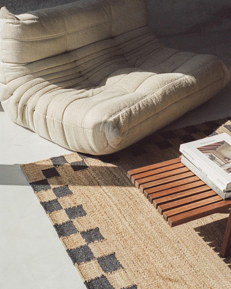 Pu&ograve; includere: Una poltrona bassa beige con tessuto strutturato, vicino a un tappeto tessuto con un motivo a scacchiera nero e beige. &Egrave; visibile anche un tavolino in legno con un libro sopra.