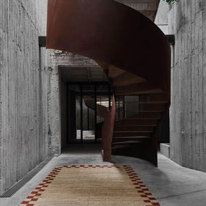 K&ouml;nnte beinhalten: Ein rechteckiger beiger Teppich mit einem rot karierten Rand liegt auf einem Betonboden. Eine Wendeltreppe mit einem rostbraunen Metall&auml;u&szlig;eren und Holzstufen windet sich nach oben. Die W&auml;nde bestehen aus strukturiertem Beton.
