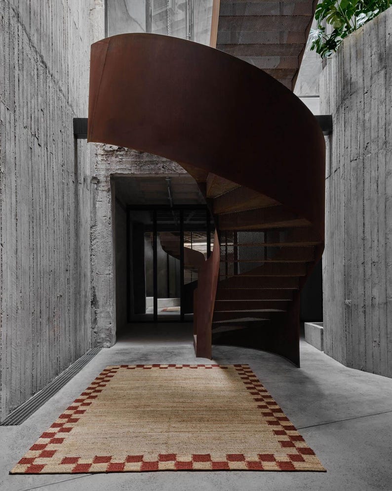 K&ouml;nnte beinhalten: Ein rechteckiger beiger Teppich mit einem rot karierten Rand liegt auf einem Betonboden. Eine Wendeltreppe mit einem rostbraunen Metall&auml;u&szlig;eren und Holzstufen windet sich nach oben. Die W&auml;nde bestehen aus strukturiertem Beton.