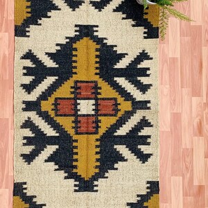 May include: A rectangular rug with a geometric pattern in cream, black, mustard yellow, and rust. The rug has a fringed edge and a central diamond design. A potted plant is in the upper right corner.