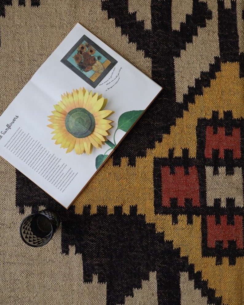May include: A still-life image featuring a book with a sunflower illustration and a 3D paper sunflower. A glass with a geometric pattern sits near the book. The background is a woven rug with black, tan, yellow, and red geometric patterns.