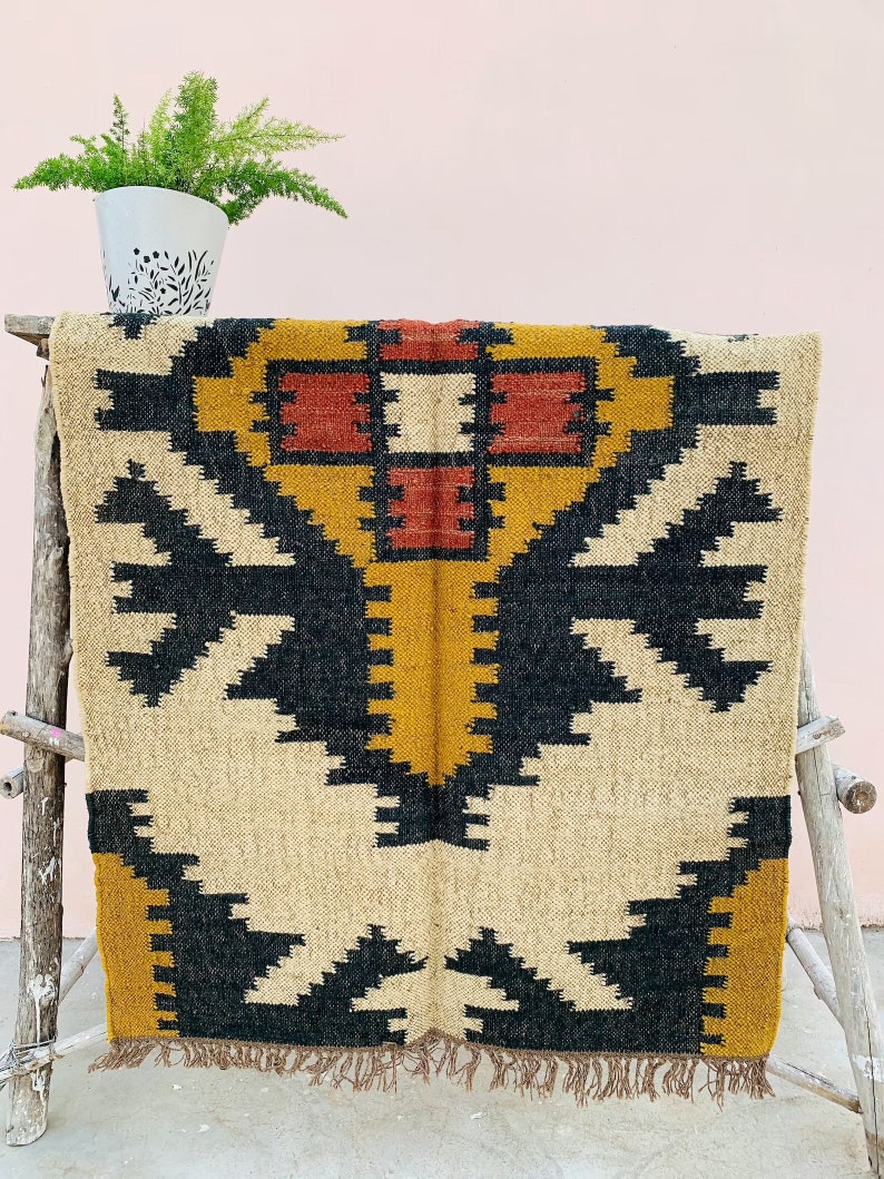 May include: A woven rug with a geometric pattern in black, mustard yellow, red, and beige. The rug is displayed on a wooden rack, with a potted plant in a white pot with a black floral design in the background. The rug has a fringed edge.