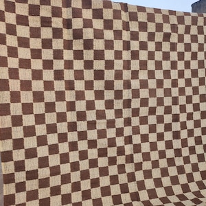 May include: A large, woven textile featuring a brown and beige chequerboard pattern. The fabric is draped, displaying the repeating square design. The textile appears to be made of a natural fibre, suitable for use as a rug or blanket.