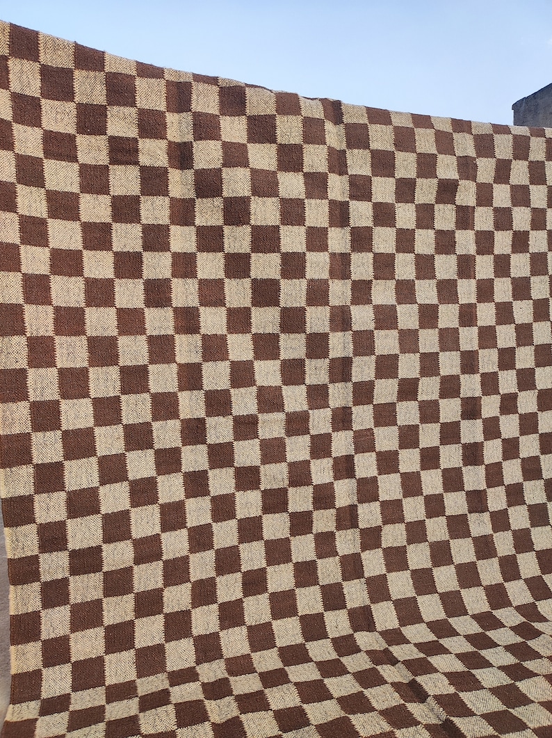 May include: A large, woven textile featuring a brown and beige chequerboard pattern. The fabric is draped, displaying the repeating square design. The textile appears to be made of a natural fibre, suitable for use as a rug or blanket.