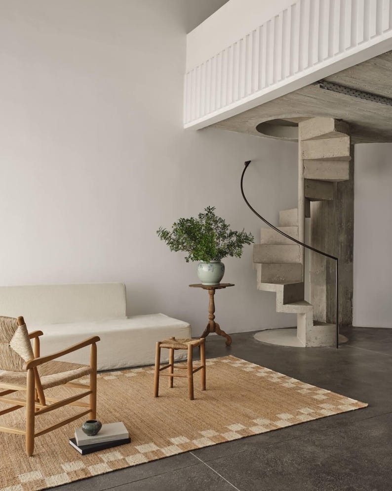 Pu&ograve; includere: Interno moderno con un tappeto beige con bordo geometrico su un pavimento scuro. Sono visibili una sedia e uno sgabello in legno, un divano bianco e un tavolino con una pianta. Una scala a chiocciola in cemento e una parete bianca completano la scena.