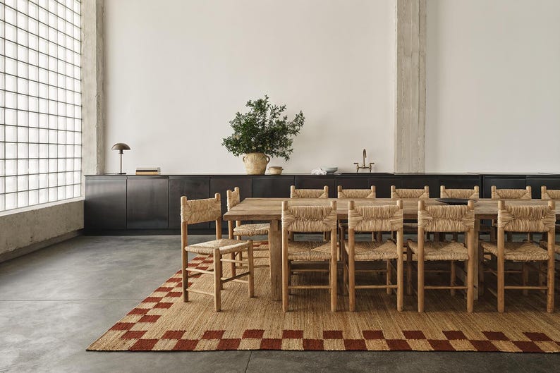 K&ouml;nnte beinhalten: Ein Esszimmer-Set mit einem Holztisch und St&uuml;hlen mit geflochtenen R&uuml;ckenlehnen. Unter dem Tisch liegt ein rechteckiger Teppich mit einem rot-beigen Karomuster. Eine gro&szlig;e Pflanze in einem geflochtenen Topf steht auf einem dunklen Schrank.