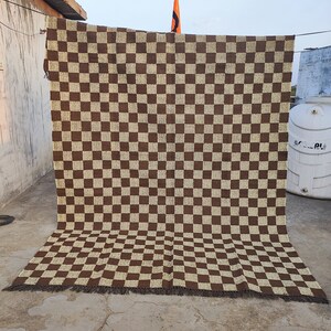 May include: A large, rectangular brown and beige chequered rug. The rug is hanging outside, with the top edge attached to a line. The rug has a fringe along the bottom edge. The pattern is a repeating grid of squares.