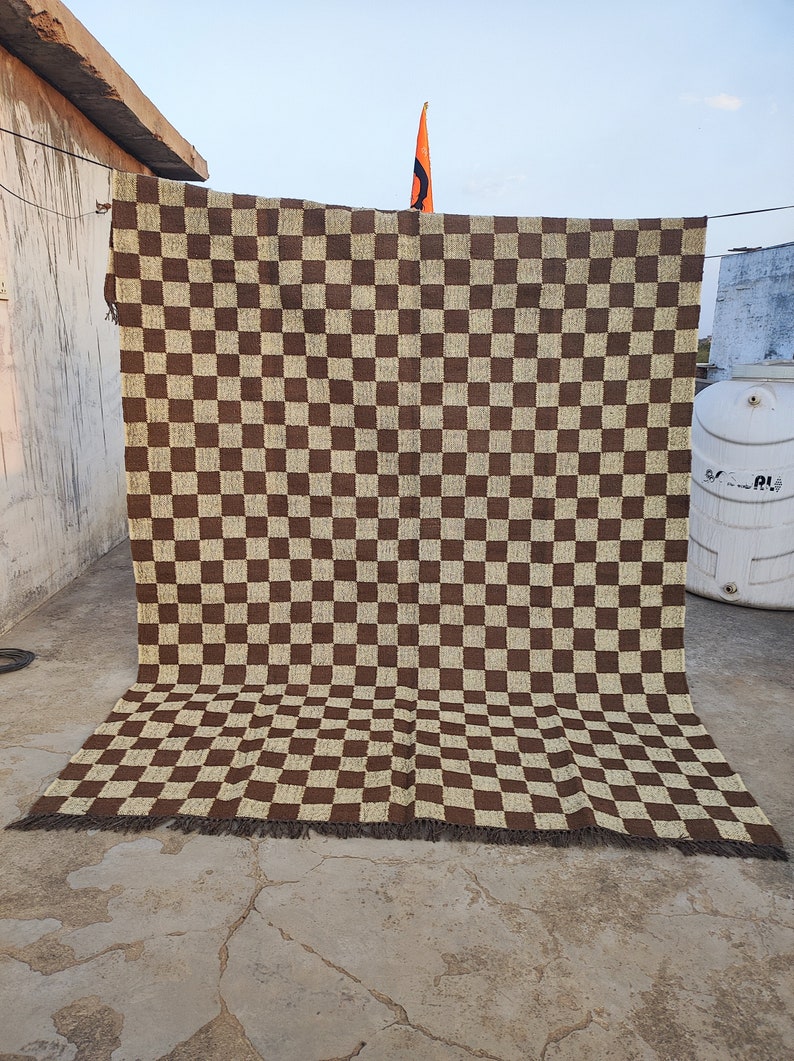 May include: A large, rectangular brown and beige chequered rug. The rug is hanging outside, with the top edge attached to a line. The rug has a fringe along the bottom edge. The pattern is a repeating grid of squares.
