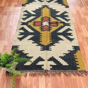 May include: A rectangular rug with a geometric pattern in black, gold, and cream. The rug has a central design with a cross-like shape, surrounded by repeating arrow-like motifs. The edges are finished with a fringe.
