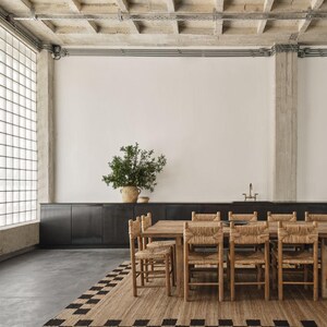 K&ouml;nnte beinhalten: Ein Esszimmer mit einem Holztisch und St&uuml;hlen mit gewebten Sitzen. Ein rechteckiger Teppich mit einem schwarz-beigen Schachbrettmuster liegt unter dem Tisch. Eine gro&szlig;e Pflanze in einem gewebten Topf.