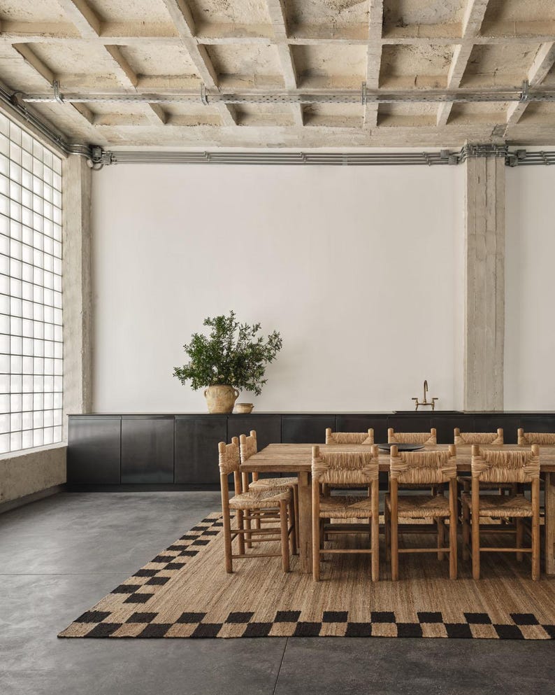 K&ouml;nnte beinhalten: Ein Esszimmer mit einem Holztisch und St&uuml;hlen mit gewebten Sitzen. Ein rechteckiger Teppich mit einem schwarz-beigen Schachbrettmuster liegt unter dem Tisch. Eine gro&szlig;e Pflanze in einem gewebten Topf.