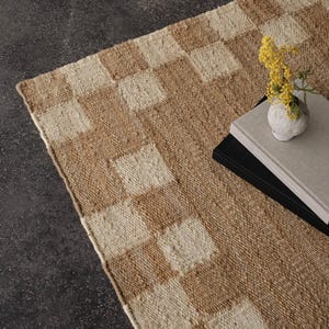 Pu&ograve; includere: Un tappeto rettangolare tessuto con un motivo a scacchiera nei toni del beige e del crema. Un piccolo vaso bianco con fiori gialli &egrave; appoggiato su una pila di libri sul tappeto. Il tappeto ha un aspetto strutturato.
