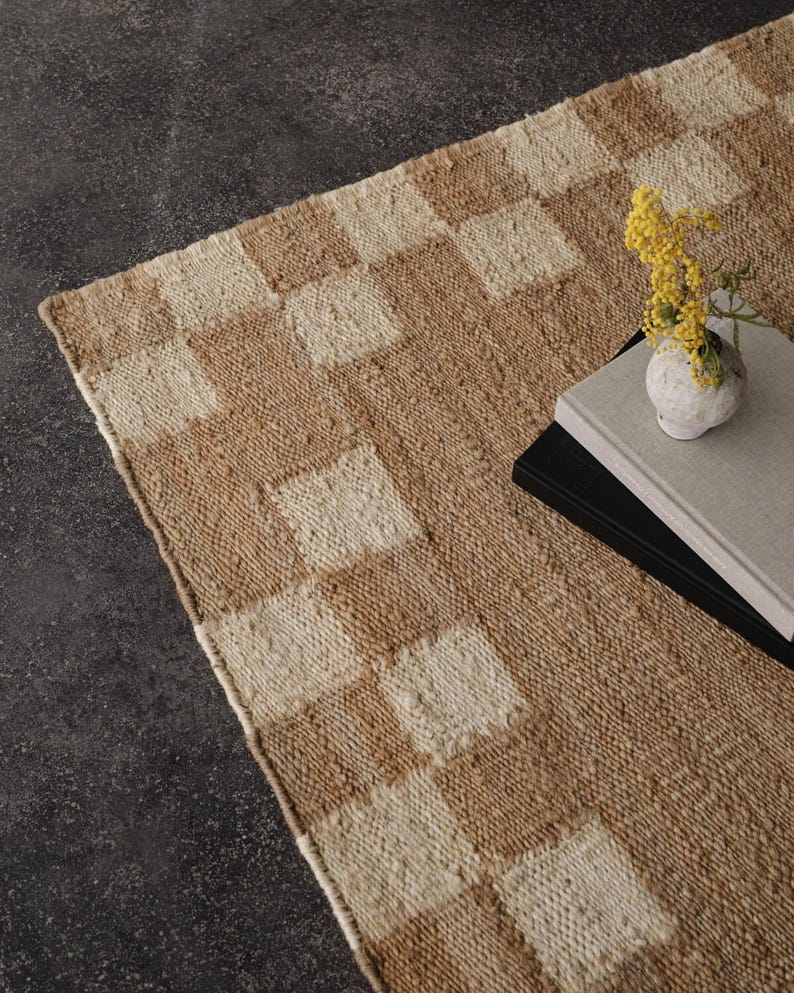 Pu&ograve; includere: Un tappeto rettangolare tessuto con un motivo a scacchiera nei toni del beige e del crema. Un piccolo vaso bianco con fiori gialli &egrave; appoggiato su una pila di libri sul tappeto. Il tappeto ha un aspetto strutturato.