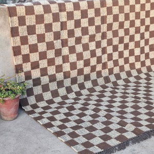 May include: A brown and beige checkered rug with a fringe edge. The rug has a large checkerboard pattern. A woven rug with a potted plant to the left. The rug's dimensions are in centimetres.