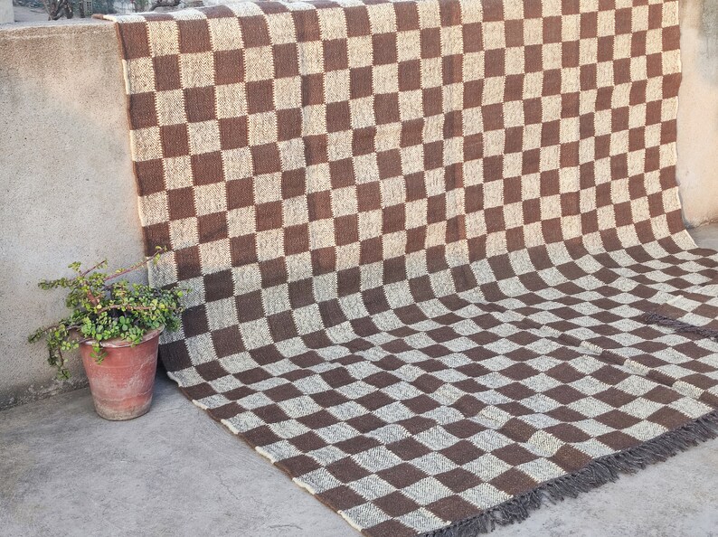 May include: A brown and beige checkered rug with a fringe edge. The rug has a large checkerboard pattern. A woven rug with a potted plant to the left. The rug's dimensions are in centimetres.
