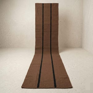 May include: A long, brown runner rug with three vertical black stripes. The rug is made of a woven material and is displayed against a neutral background. The runner is a decorative home decor item.