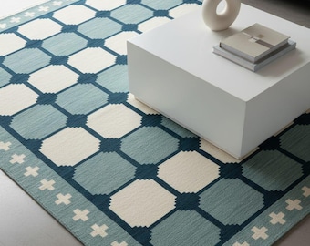 Könnte beinhalten: Rechteckiger Teppich mit geometrischem Muster in Blau- und Cremetönen. Der Teppich hat ein sich wiederholendes quadratisches Design mit einem Rand aus kreuzförmigen Akzenten. Ein weißer Couchtisch und ein graues Sofa befinden sich im Hintergrund.