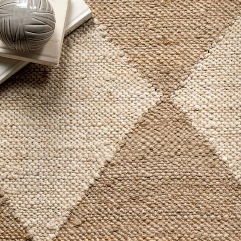 May include: Close-up of a textured, neutral-toned rug with a geometric pattern. The rug features diagonal sections of light beige and medium brown, creating a textured surface. A decorative gray ball and books are visible in the upper left corner.