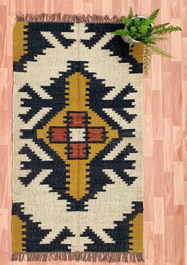 May include: Rectangular rug with a geometric pattern in cream, black, mustard yellow, and rust. The rug has a fringed edge and a central diamond-like design. A small potted plant with green foliage is in the upper right corner.