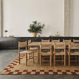 Pu&ograve; includere: Una sala da pranzo con un tavolo in legno e sedie con schienale intrecciato. Un tappeto rettangolare con un motivo a scacchi rossi e beige &egrave; sotto il tavolo. Una grande pianta in un vaso intrecciato &egrave; su un mobile scuro.