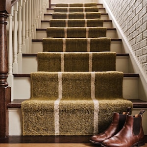May include: A staircase with olive green carpet runners featuring vertical cream stripes. Brown leather boots sit on the bottom step. The stairs have wooden steps, white risers, and a wooden handrail.