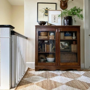 May include: A vintage wooden cabinet with glass doors, filled with books and decorative items, sits on a diamond-patterned rug. Above the cabinet are framed insect prints, a decorative bowl, and a deer antler mount. The room has a warm, inviting atmosphere.