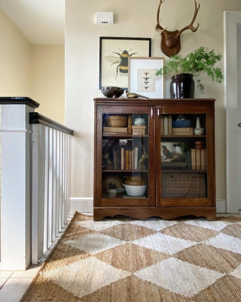 May include: A vintage wooden cabinet with glass doors, filled with books and decorative items, sits on a diamond-patterned rug. Above the cabinet are framed insect prints, a decorative bowl, and a deer antler mount. The room has a warm, inviting atmosphere.