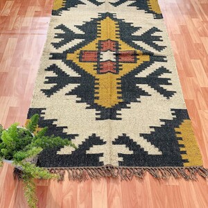 May include: A rectangular rug with a geometric pattern in black, gold, and cream. The rug has a central design with a cross-like shape, surrounded by repeating arrow-like motifs. The edges are finished with a fringe.