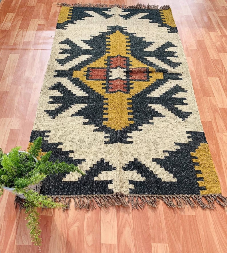 May include: A rectangular rug with a geometric pattern in black, gold, and cream. The rug has a central design with a cross-like shape, surrounded by repeating arrow-like motifs. The edges are finished with a fringe.