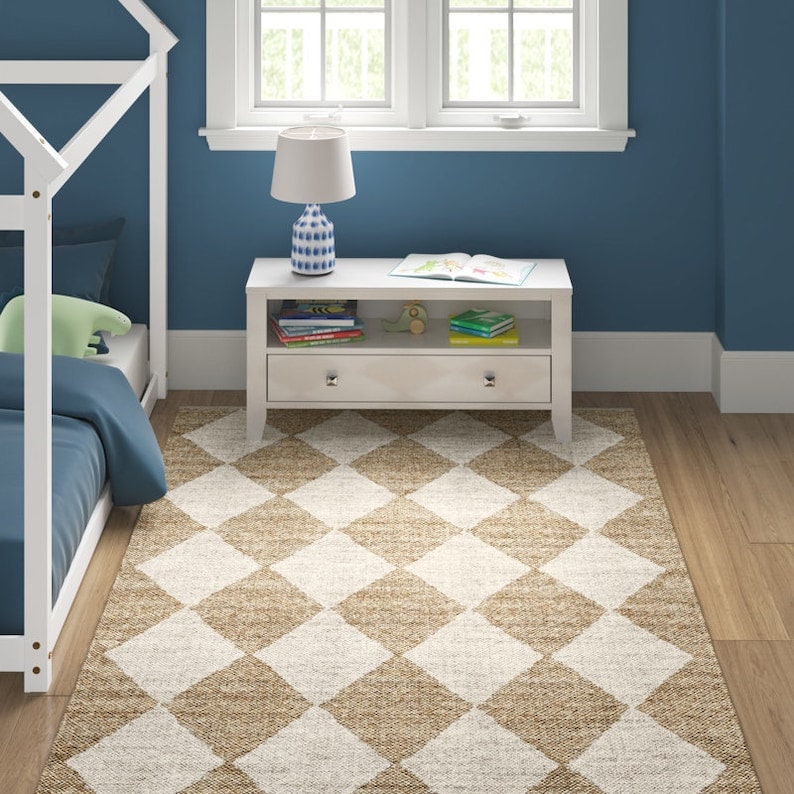 May include: A rectangular area rug with a diamond pattern in beige and off-white. The rug is placed on a hardwood floor in a child's bedroom. A white nightstand with a blue and white lamp sits next to a bed.