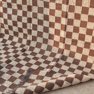 May include: A brown and beige checkered woven rug. The rug features a repeating pattern of squares in two colours. The rug has a fringed edge on one side. The rug is made of a thick, textured material.