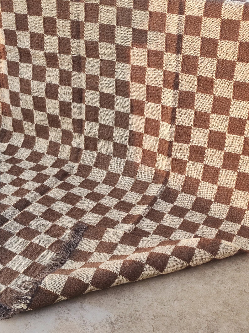 May include: A brown and beige checkered woven rug. The rug features a repeating pattern of squares in two colours. The rug has a fringed edge on one side. The rug is made of a thick, textured material.