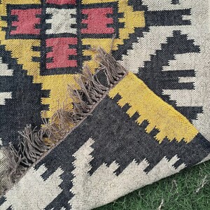 May include: A woven rug with a geometric pattern in black, yellow, white, and red. The rug has a fringed edge and is partially folded, revealing the underside. The design features repeating shapes and a central square motif.