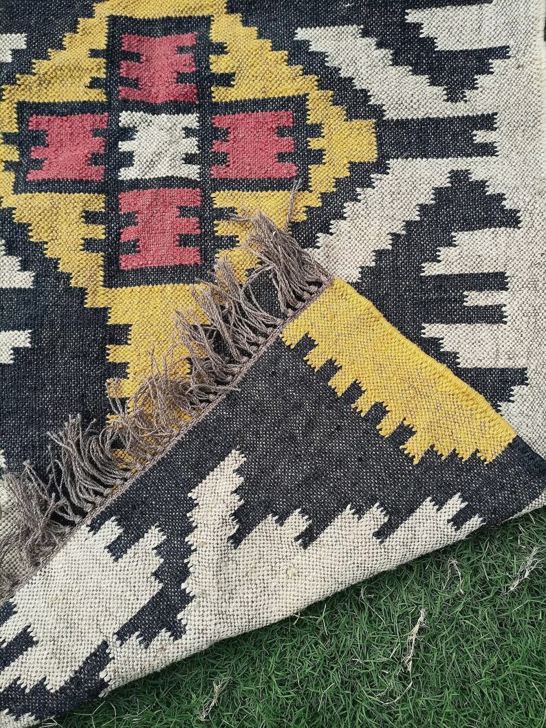May include: A woven rug with a geometric pattern in black, yellow, white, and red. The rug has a fringed edge and is partially folded, revealing the underside. The design features repeating shapes and a central square motif.
