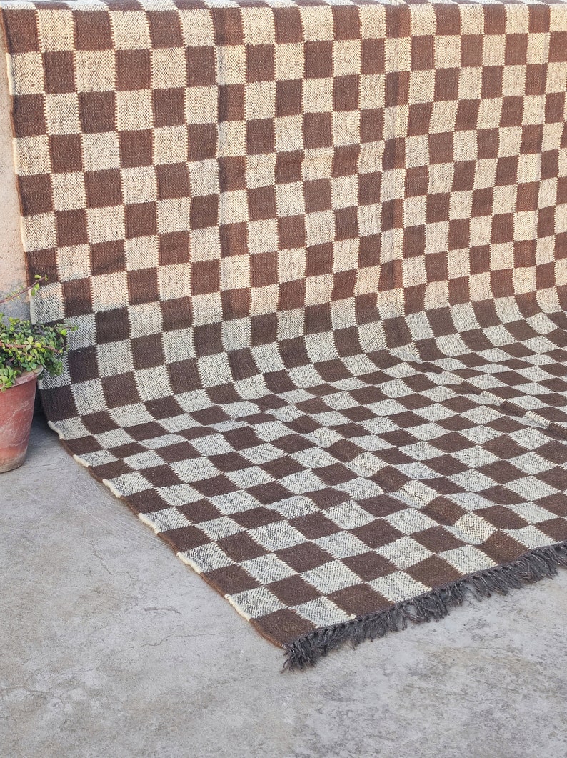May include: A brown and white checkered rug with fringe on the bottom edge. The rug is made of a woven material and has a slightly textured surface.