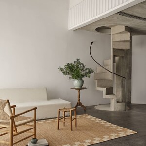K&ouml;nnte beinhalten: Ein minimalistischer Wohnraum mit beigem Teppich und geometrischem Rand. Ein wei&szlig;es Sofa, ein Holzstuhl und ein Hocker sind auf dem Teppich angeordnet. Ein kleiner Beistelltisch tr&auml;gt eine Vase mit Gr&uuml;n. Eine Wendeltreppe aus Beton f&uuml;hrt in eine zweite Ebene.