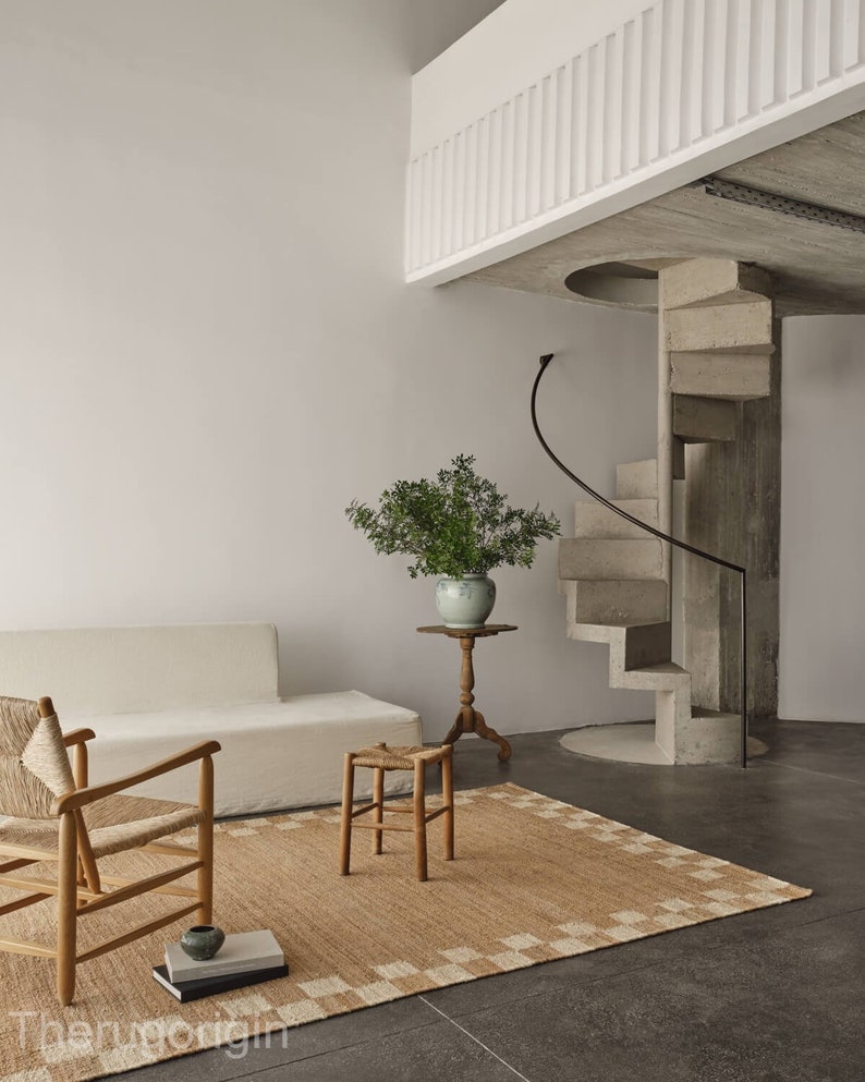 K&ouml;nnte beinhalten: Ein minimalistischer Wohnraum mit beigem Teppich und geometrischem Rand. Ein wei&szlig;es Sofa, ein Holzstuhl und ein Hocker sind auf dem Teppich angeordnet. Ein kleiner Beistelltisch tr&auml;gt eine Vase mit Gr&uuml;n. Eine Wendeltreppe aus Beton f&uuml;hrt in eine zweite Ebene.