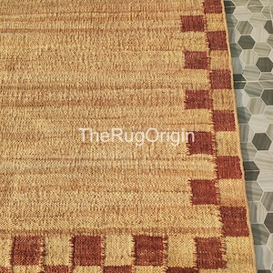 Pu&ograve; includere: Un tappeto rettangolare in juta con una trama intrecciata e un bordo di quadrati marroni e beige alternati. Il tappeto ha una palette di colori naturali e il testo "TheRugOrigin" &egrave; visibile.