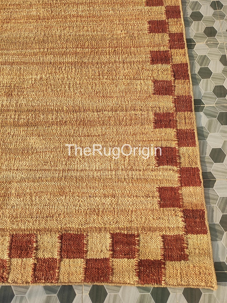 Pu&ograve; includere: Un tappeto rettangolare in juta con una trama intrecciata e un bordo di quadrati marroni e beige alternati. Il tappeto ha una palette di colori naturali e il testo "TheRugOrigin" &egrave; visibile.