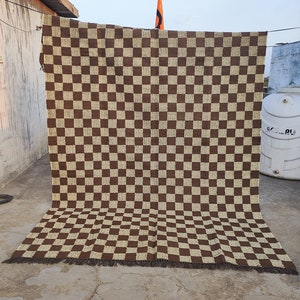 May include: A large, rectangular brown and beige chequered rug with a fringe along the bottom edge. The rug is hanging outside, with a flag visible in the background. The pattern is a repeating grid of squares.