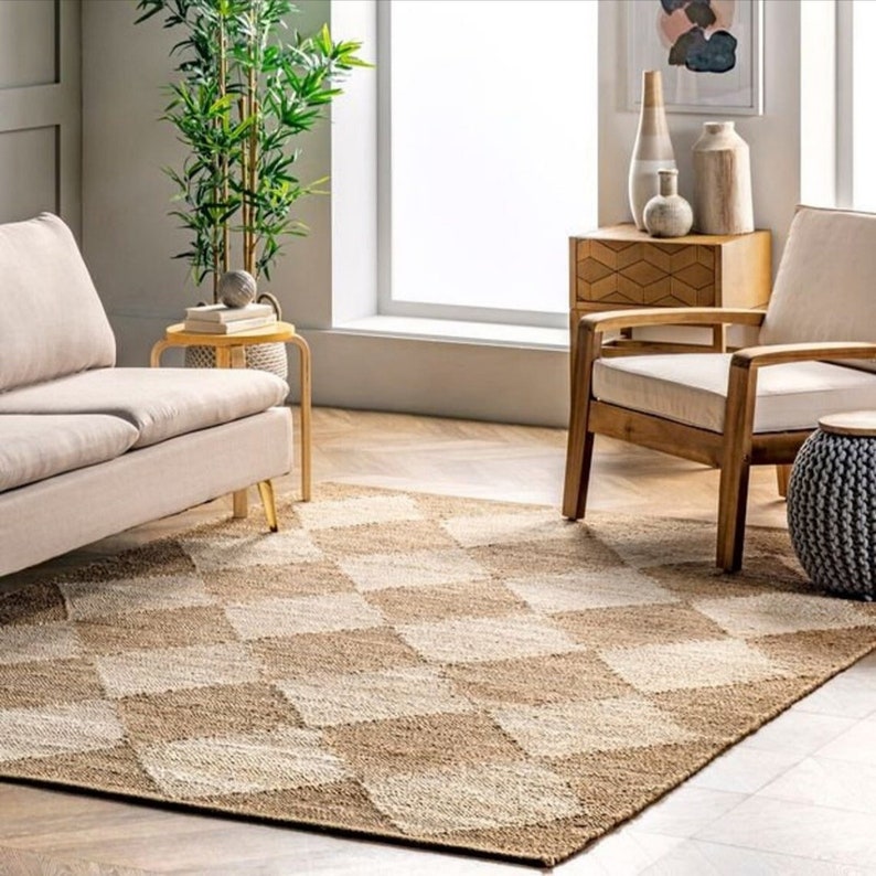 May include: A neutral-toned living room featuring a geometric patterned rug in shades of beige and tan. The rug is the focal point, complemented by a beige sofa, wooden chairs, and decorative vases. A potted plant adds a touch of greenery.