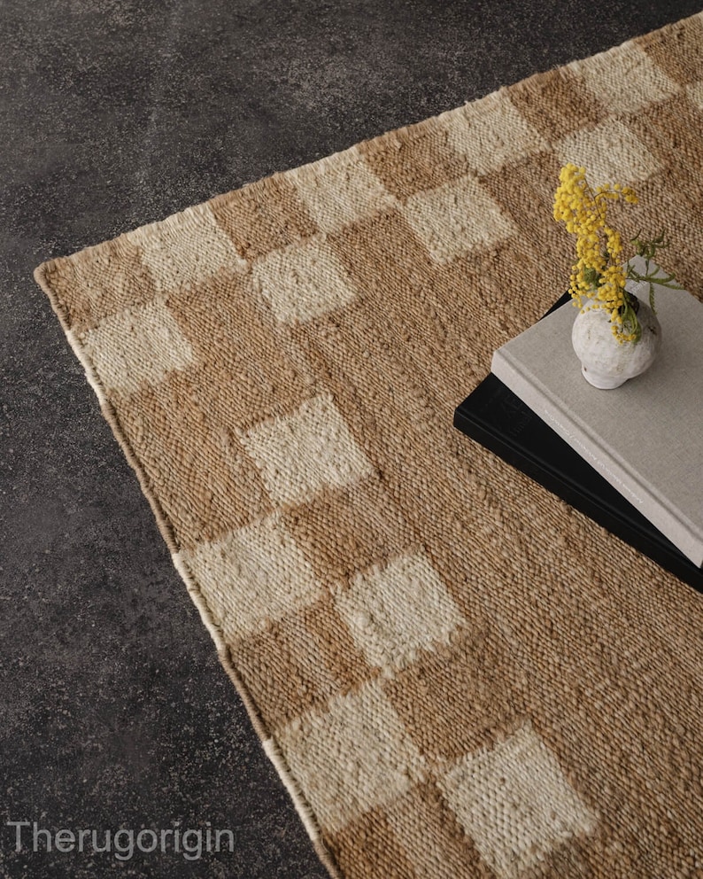 K&ouml;nnte beinhalten: Ein rechteckiger, gewebter Teppich mit einem Schachbrettmuster in Beige- und Braunt&ouml;nen. Der Teppich liegt auf einer dunklen, strukturierten Oberfl&auml;che. Eine kleine Vase mit gelben Blumen und zwei B&uuml;cher befinden sich auf der rechten Seite des Teppichs.